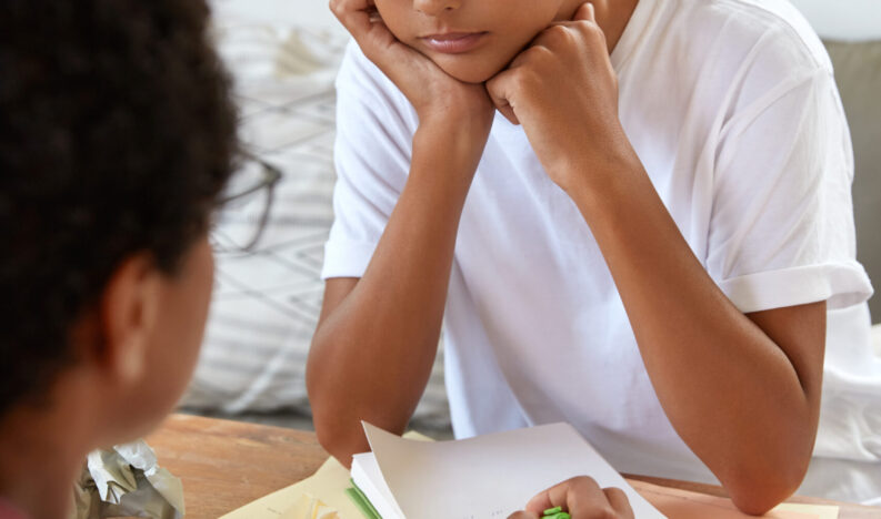 Vertical shot of Asian trainee wears spectacles and white t shirt, holds hands under chin, looks attentively at female tutor who points with pen in notebbok, suggests new strategy, helps to do task